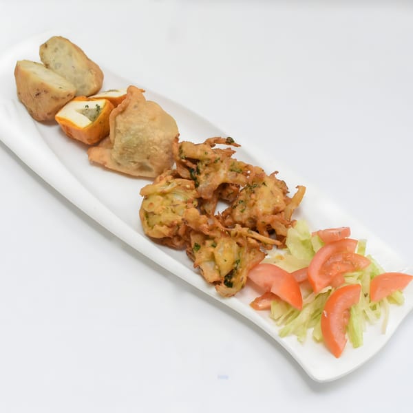 A white platter with Indian appetizers: samosas, pakoras, potato wedges, and a side salad.