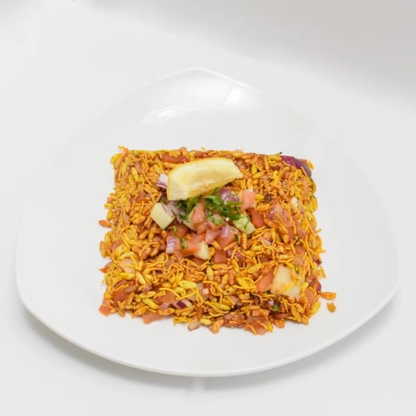 A square plate of Indian bhel puri, topped with diced vegetables and a lemon wedge.