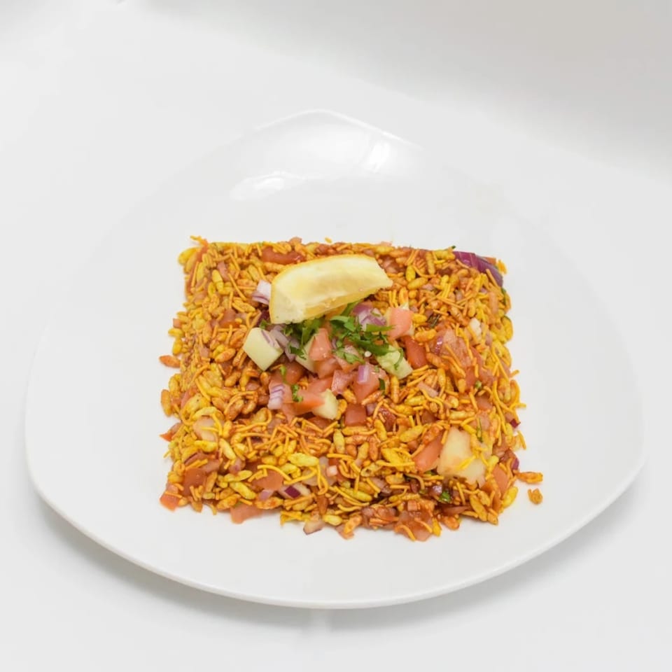 A square plate of Indian bhel puri, topped with diced vegetables and a lemon wedge.